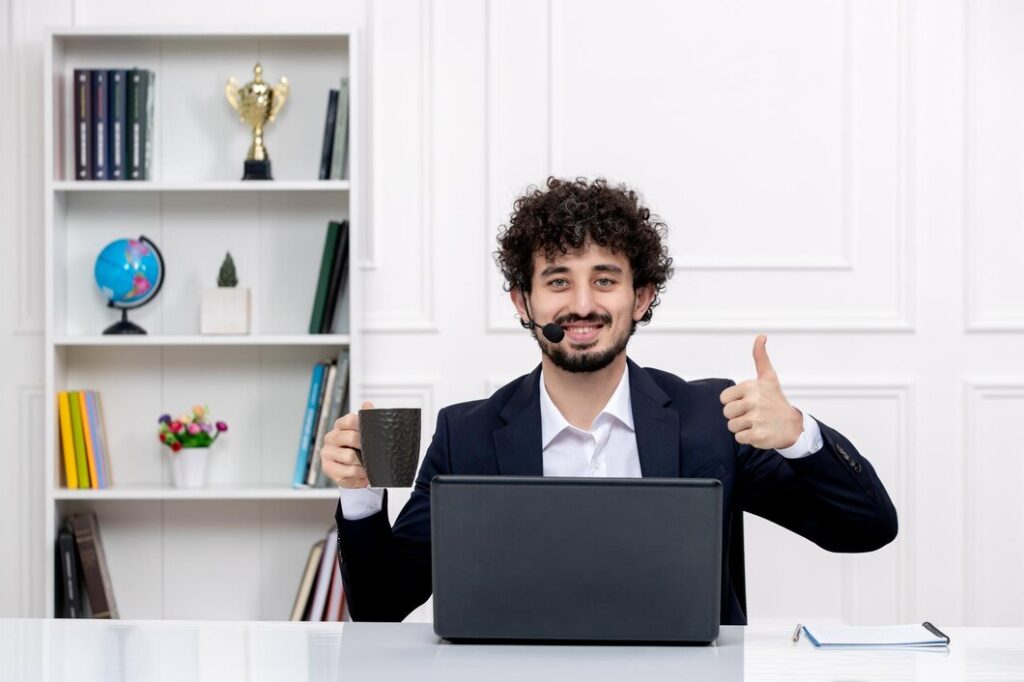 Man in a suit with a headset, holding a coffee mug and giving a thumbs-up at a desk with a laptop. Bookshelf with a trophy and globe behind him. The mood is positive and professional.
